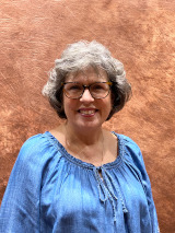 white woman with short gray wavy hair wearing glasses and a light blue blouse
