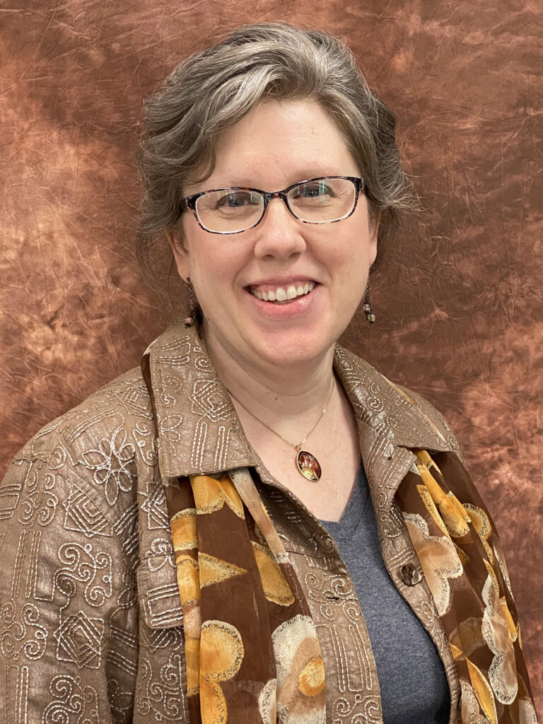White woman with short grey hair, glasses, wearing a grey shirt, tan collared jacket and brown scarf with a muted orange mushroom pattern