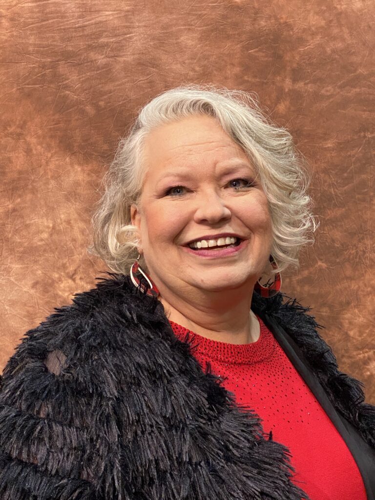 older white woman with short wavy gray hair wearing a brown fur coat and red shirt
